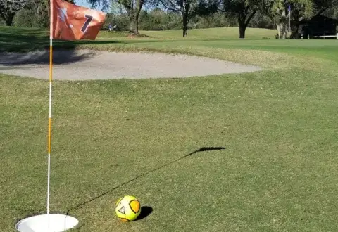 A yellow soccer ball sits near a golf hole with a flag marked 7 on a grassy course, with a sand bunker and trees in the background.