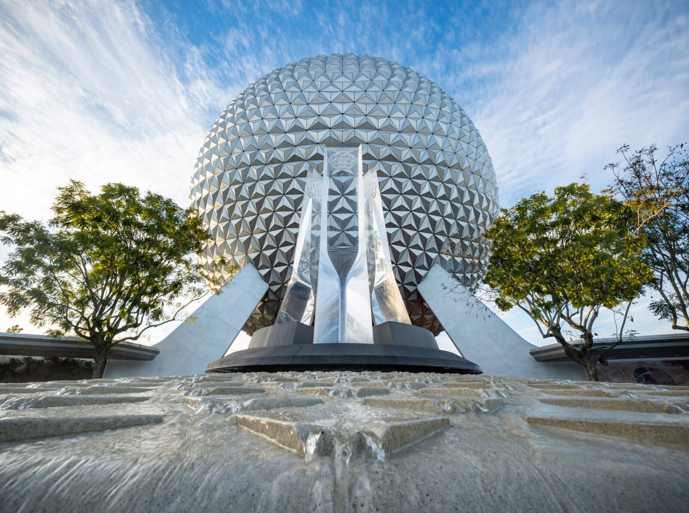 A close-up view of the iconic geodesic sphere Spaceship Earth at EPCOT in Disney World, backed by a blue sky, with water flowing in the foreground and leafy green trees on either side.