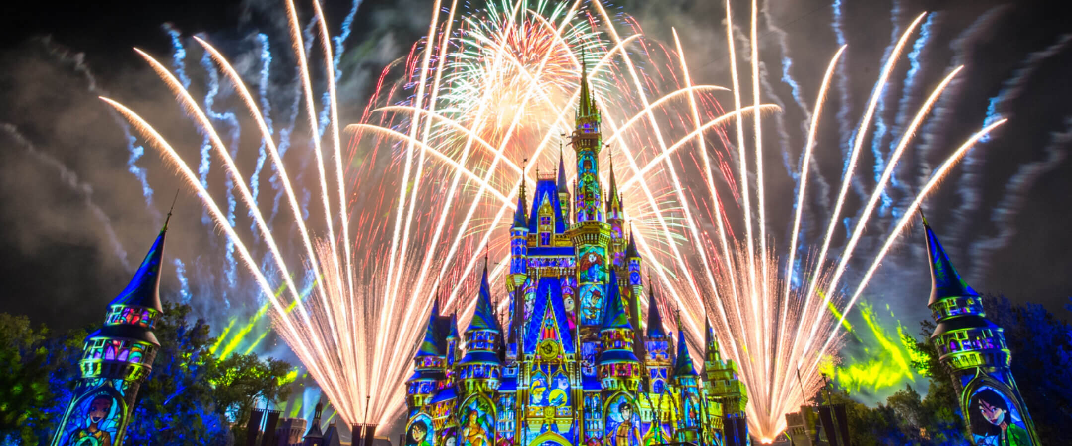 A brightly illuminated castle with colorful projections on its walls stands in the foreground, while large, vibrant fireworks light up the night sky behind it, creating a festive and magical atmosphere.