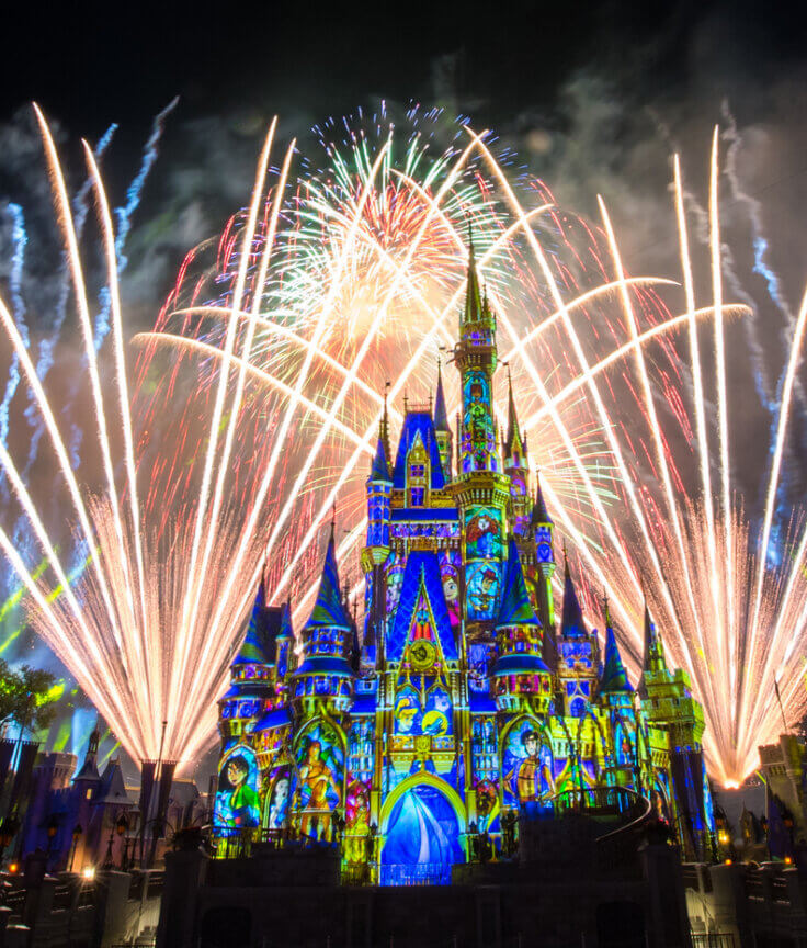 Em primeiro plano, ergue-se um castelo intensamente iluminado com projeções coloridas nas paredes, enquanto grandes e vibrantes fogos de artifício iluminam o céu noturno ao fundo, criando uma atmosfera festiva e mágica.