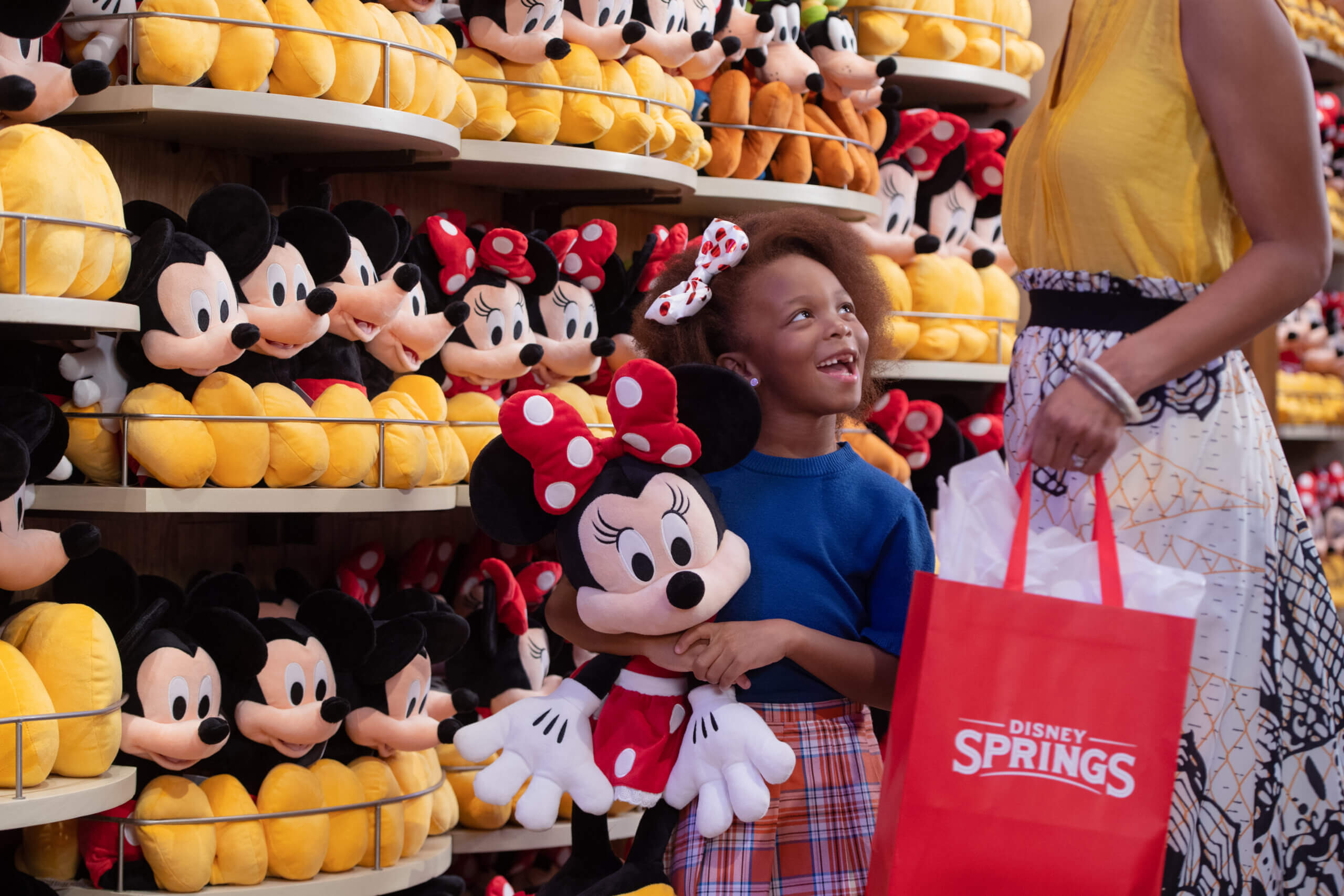 A smiling child holds a Minnie Mouse plush toy in a store filled with Mickey and Minnie Mouse stuffed animals, while an adult stands nearby holding a Disney Springs shopping bag.