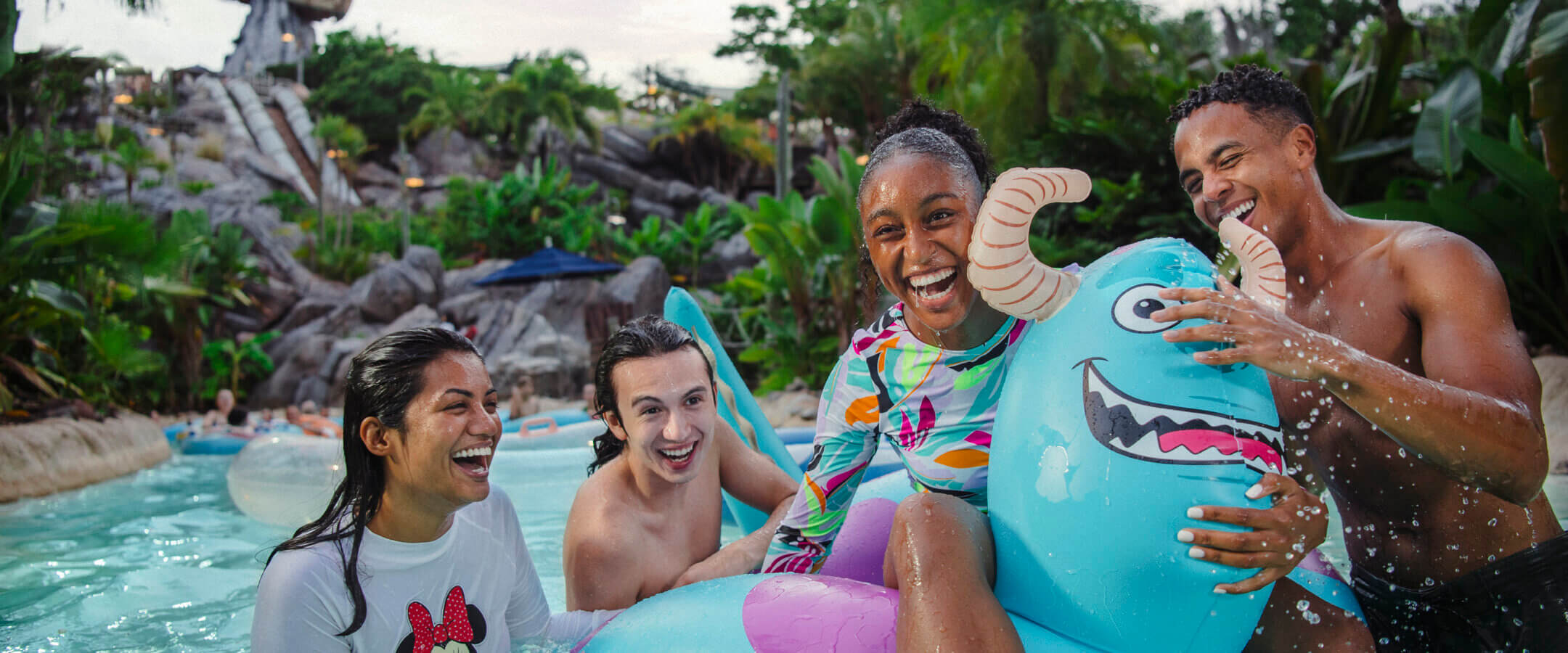 Four friends laugh and play together in a lazy river at a water park, surrounded by lush greenery. One person rides a blue monster float, and another wears a shirt with a Minnie Mouse design.