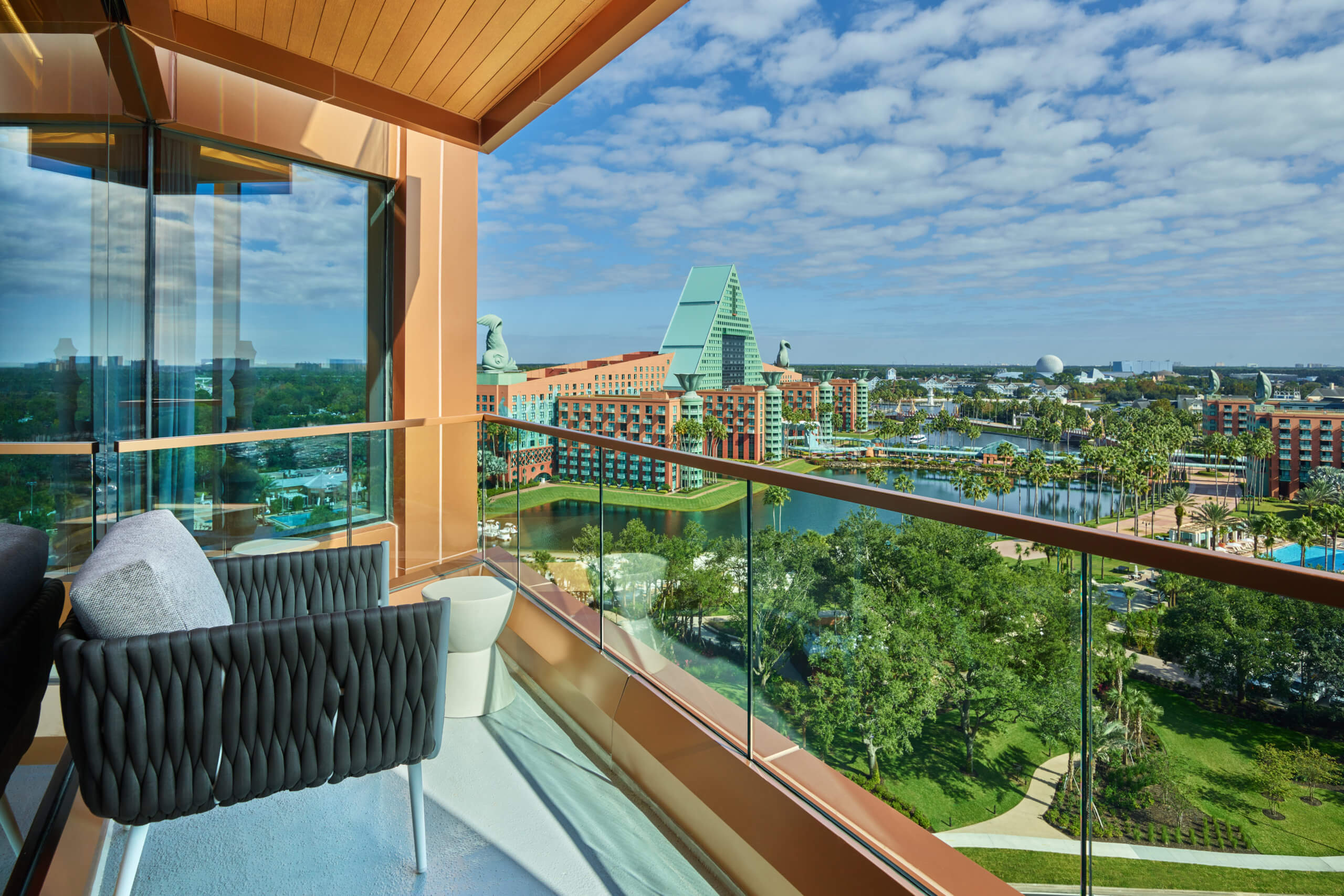 Hotel balcony with glass railing overlooks a resort with colorful buildings, water, palm trees, and green landscaping under a blue sky with clouds. A pair of modern black chairs and a small white table are in the foreground.