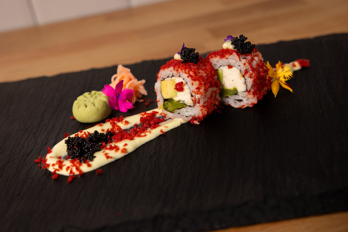 Two sushi rolls topped with red flakes and black caviar, garnished with edible flowers and wasabi, are artfully plated on a slate board with creamy sauce.