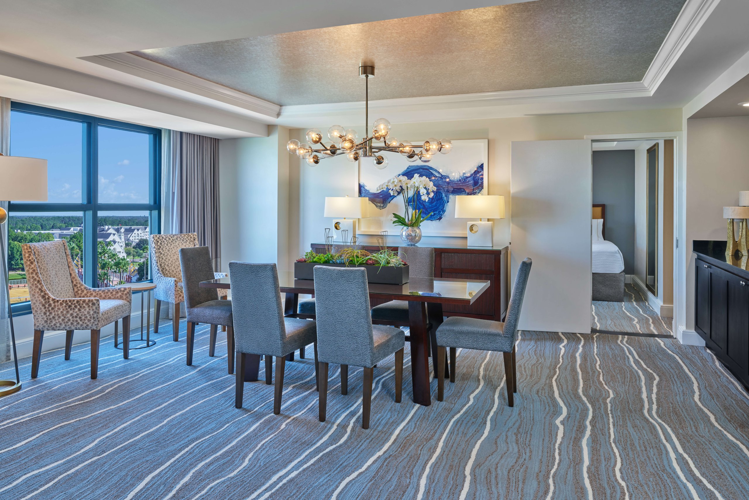 Modern dining area with six gray upholstered chairs around a dark wood table, blue abstract art on the wall, large windows, a unique chandelier, and a bedroom visible through an open doorway.