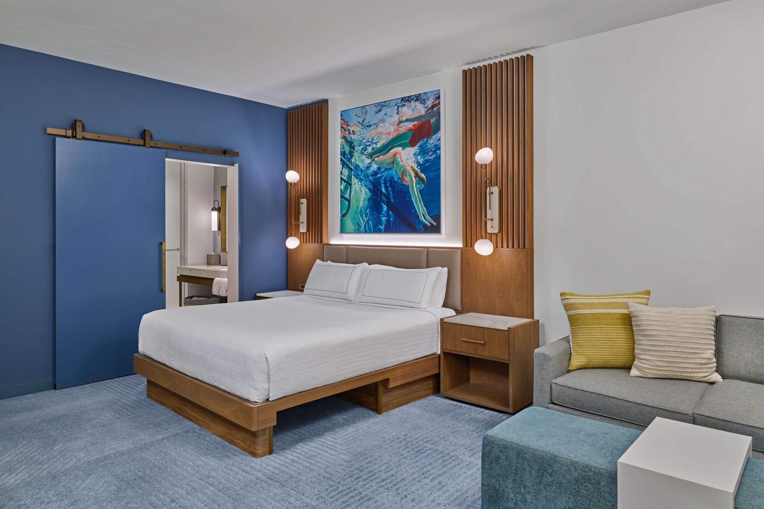 Modern hotel room with a king bed, blue walls, wood accents, a colorful underwater painting above the bed, a sliding barn door to the bathroom, and a gray sofa with yellow and white pillows.