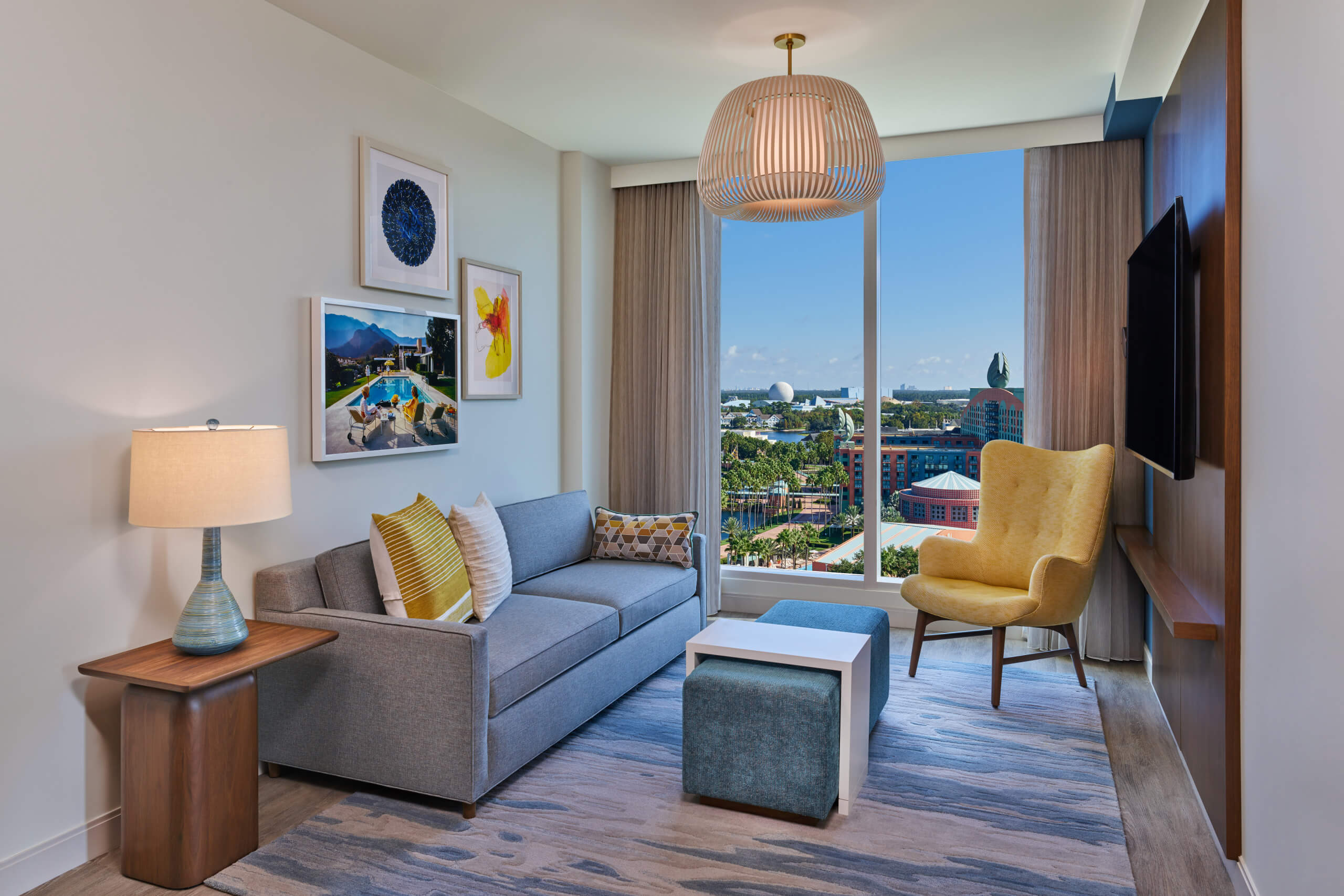 A modern living room with a gray sofa, yellow armchair, lamp, wall art, ottoman, and large window showing a cityscape with trees and blue sky.