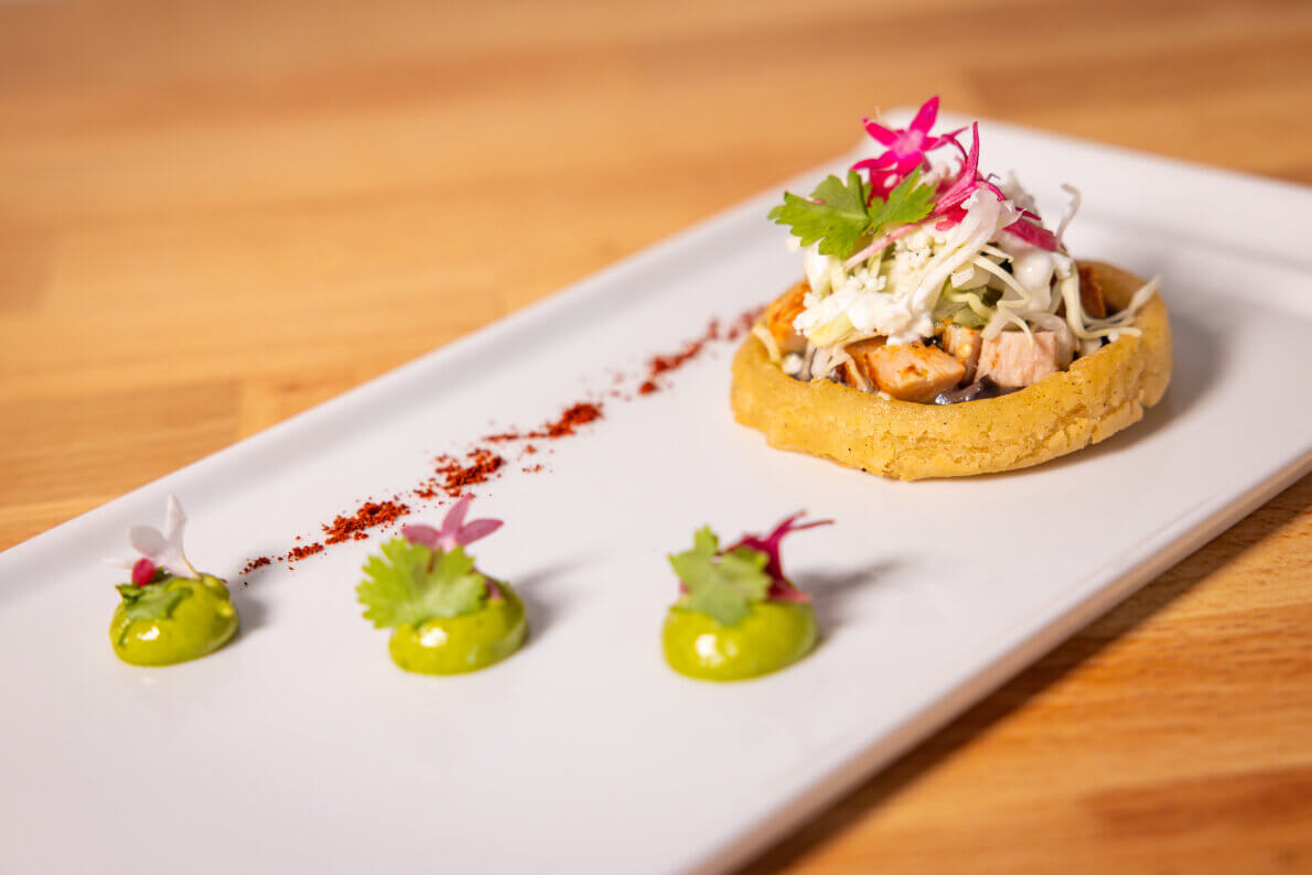 A gourmet dish with a small tostada topped with diced meat, slaw, and herbs, garnished with edible flowers. Three green sauce dollops and a line of red seasoning are artfully placed on a white plate.