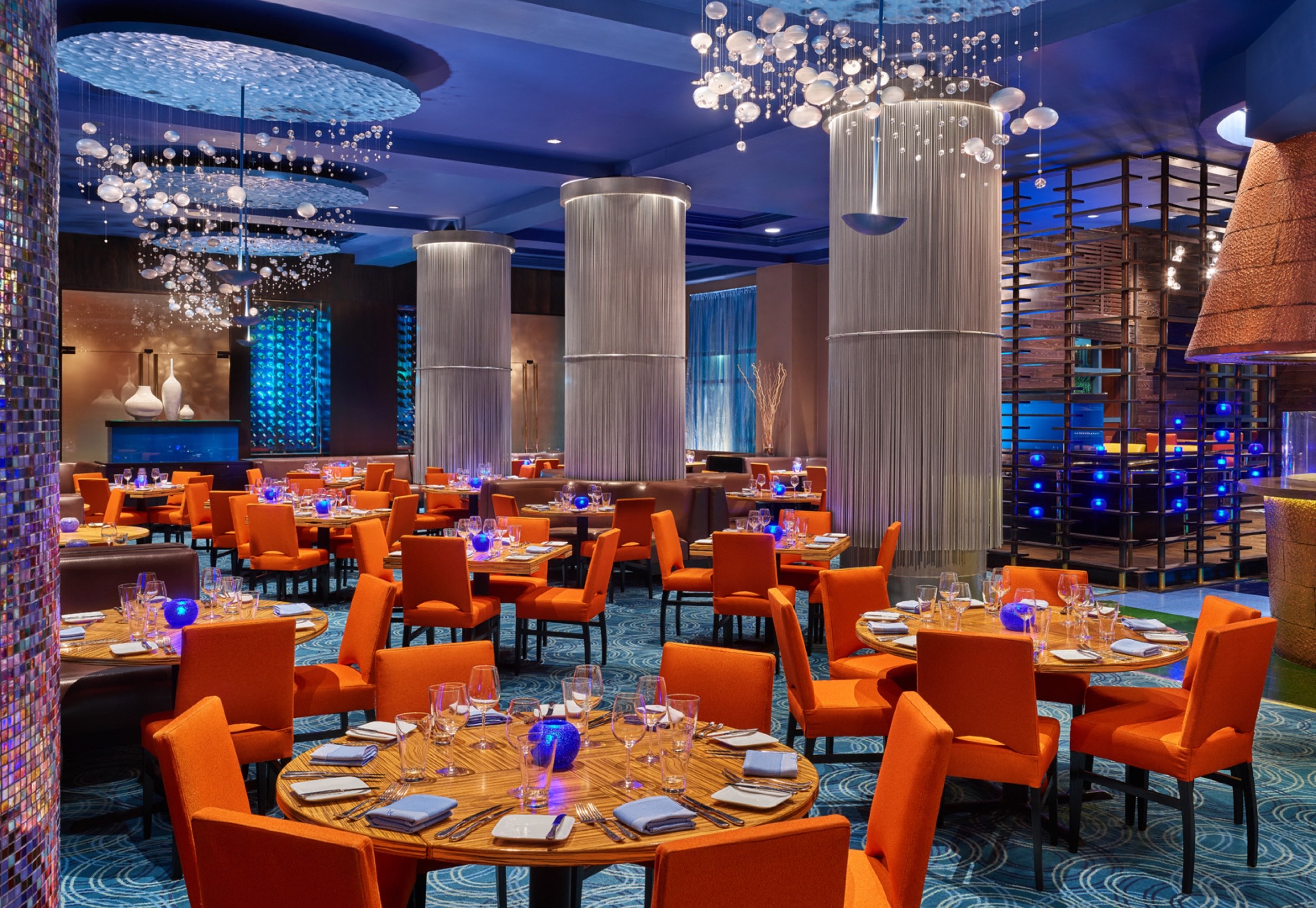 A modern restaurant with orange chairs, round wooden tables set for dining, blue lighting, tall white columns, decorative chandeliers, and a stylish, upscale ambiance.
