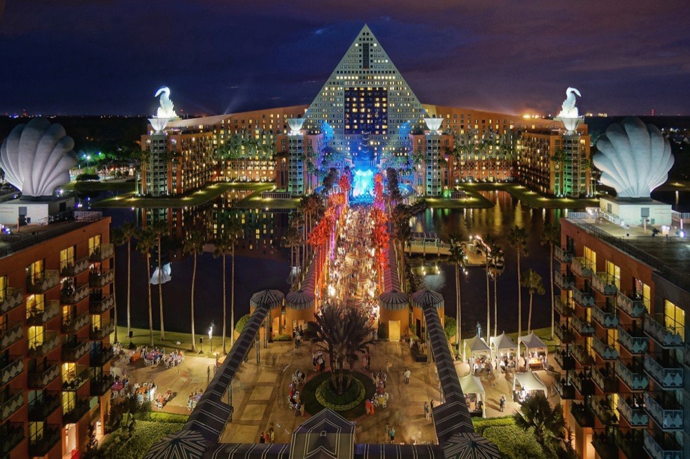 Night view of the illuminated Walt Disney World Dolphin Hotel with a pyramid shape, dolphin statues on the roof, lit palm trees, and crowds gathered along a central walkway.