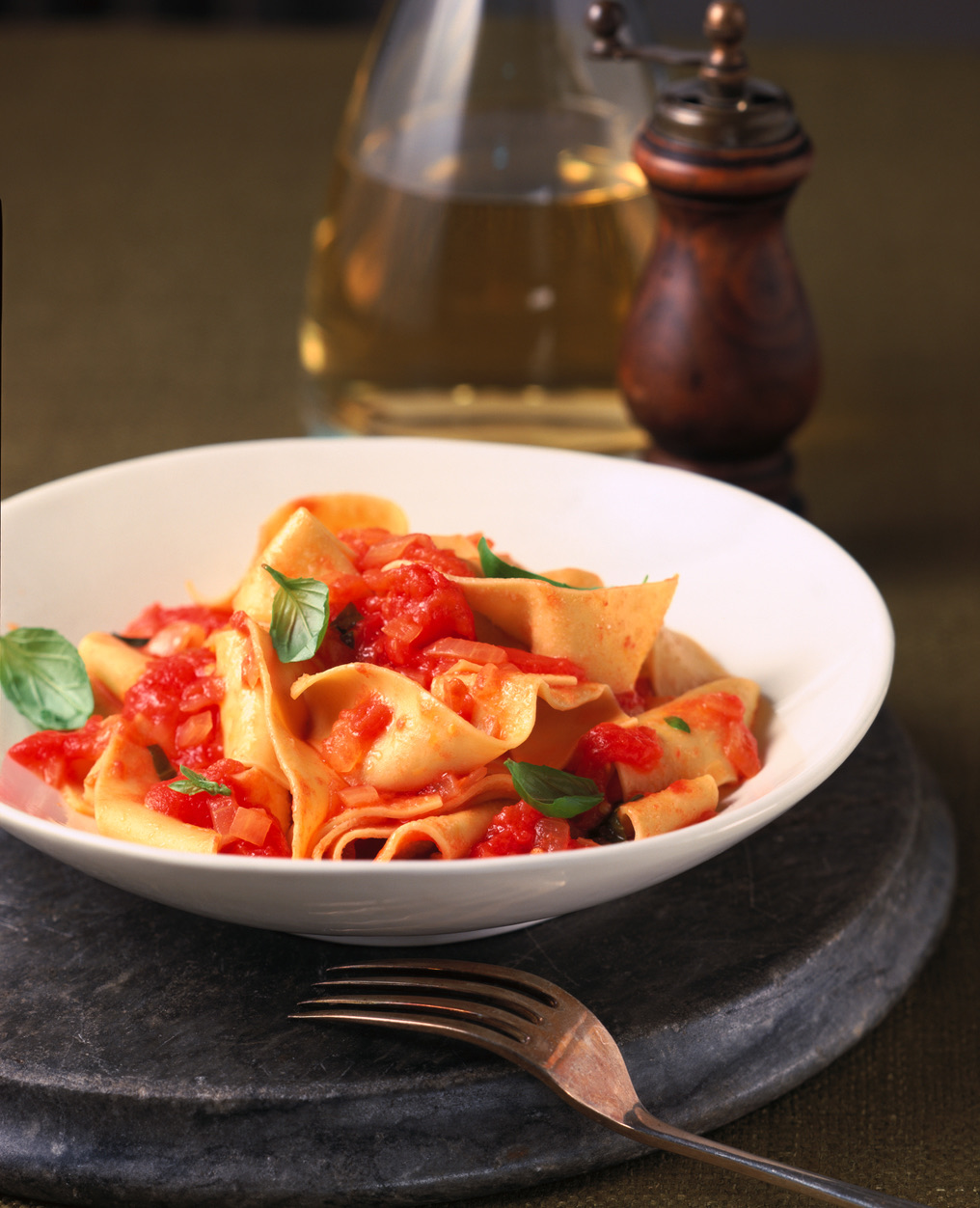 A white bowl of pappardelle pasta with tomato sauce and basil sits on a gray tray, accompanied by a fork, a glass bottle of oil, and a wooden pepper grinder in the background.