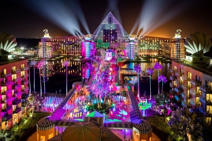 A brightly lit resort at night with a pyramid-shaped building, colorful lights, palm trees, water reflections, and beams of light shining into the sky. The scene is vibrant and festive.