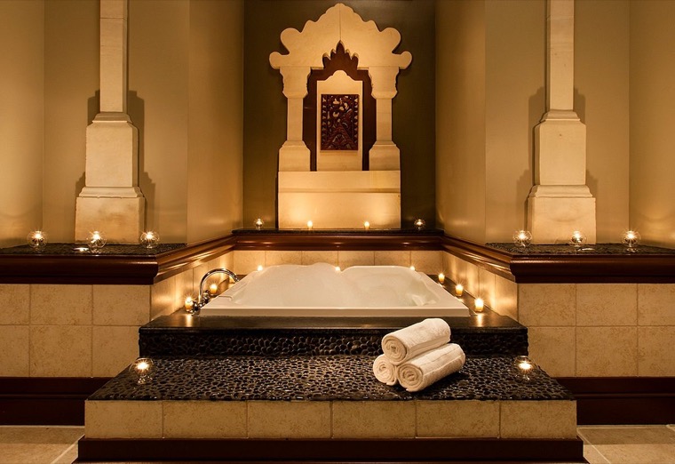 A luxurious spa bath surrounded by candles, rolled white towels, and ornate architectural details, creating a relaxing, elegant atmosphere.