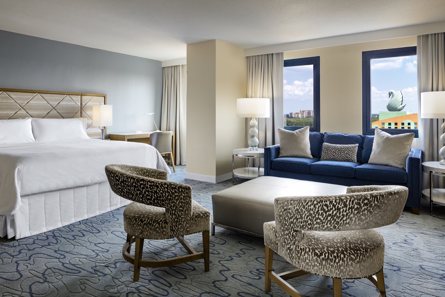 A spacious hotel suite with a king bed, two patterned chairs, a blue couch, a coffee table, and large windows letting in natural light. The decor is modern with neutral tones and soft blue accents.