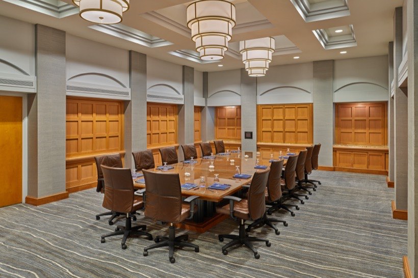 A modern conference room with a long wooden table, leather chairs, water glasses, and notepads. The room has wood-paneled walls, patterned carpet, and large rectangular ceiling lights.