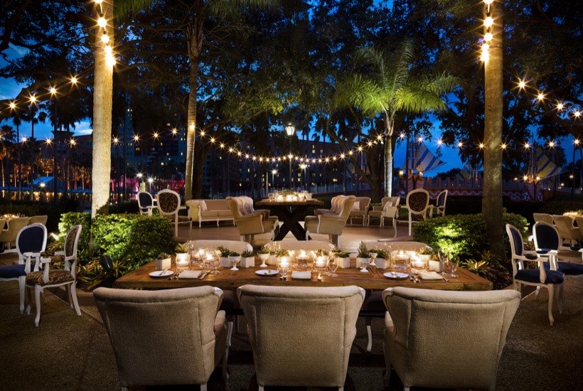 A long wooden table set for dinner outdoors, surrounded by cushioned chairs, with string lights hanging above. Lush greenery and palm trees frame the scene during twilight. Candles and small plants decorate the table.
