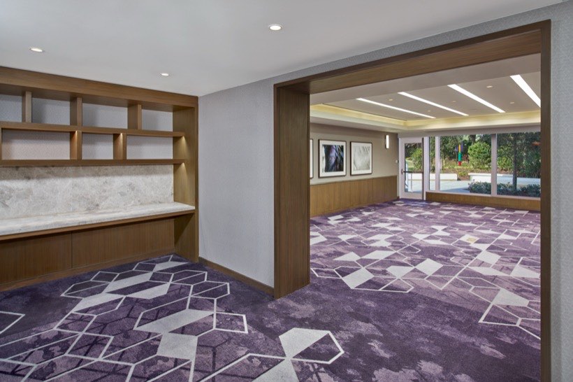 Spacious, modern room with geometric purple carpet, wooden shelves, large windows letting in natural light, and framed art on the walls.