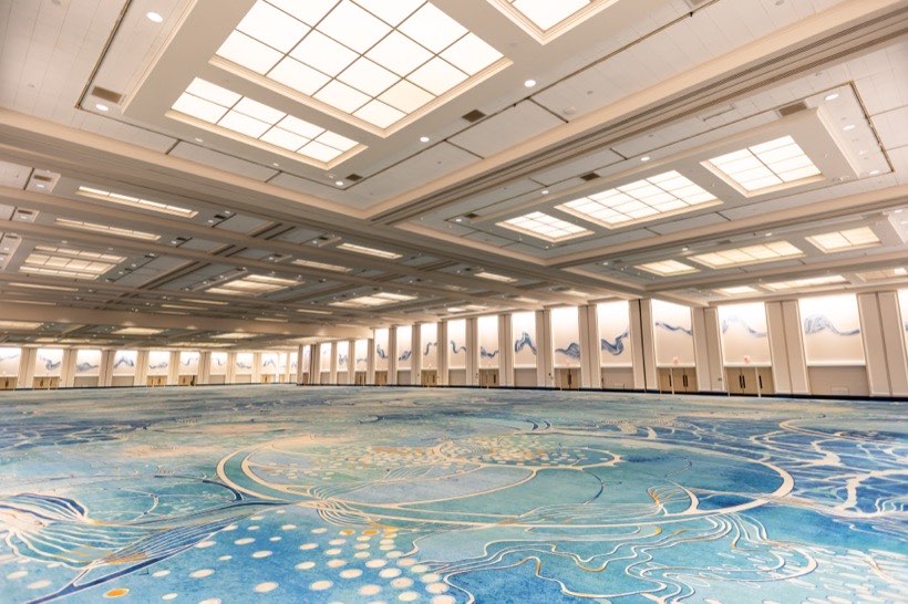 Spacious, empty conference hall with high ceilings, large ceiling lights, and a blue patterned carpet. Walls display abstract art panels.