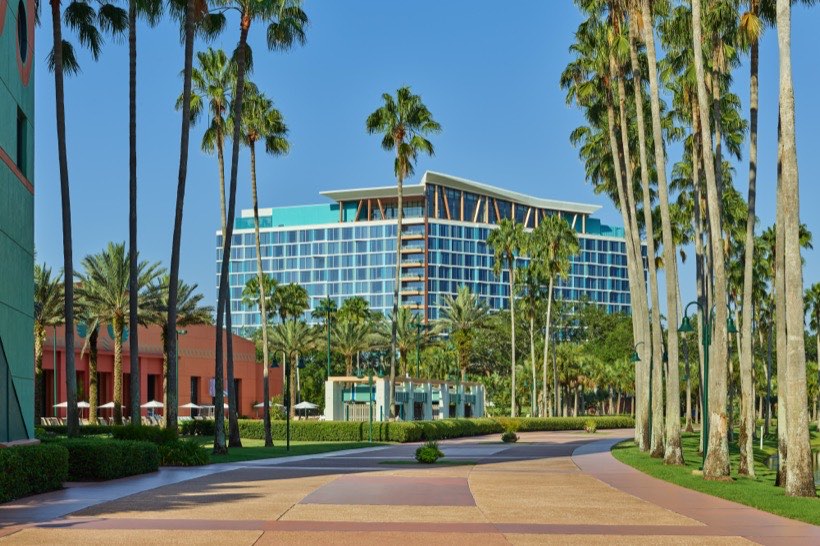 A modern, blue hotel building is surrounded by tall palm trees and lush greenery under a clear blue sky, with a wide, empty walkway in the foreground.