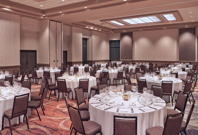 Large banquet hall with round tables set for a formal event. Each table is covered with a white tablecloth and surrounded by brown chairs. The room features neutral decor, carpet, and high ceilings with overhead lighting.