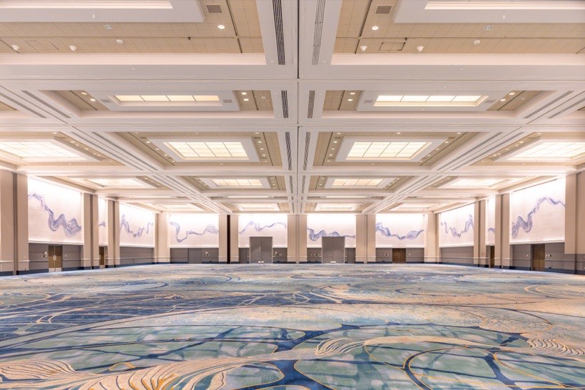 A large, empty convention hall with high ceilings, patterned carpet in blue tones, wall art with abstract blue lines, and bright ceiling lights. Multiple doors line the walls.