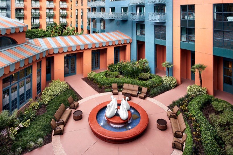 Outdoor courtyard with orange and blue buildings, circular fountain with white swan sculptures in the center, surrounded by green bushes and brown cushioned seating.