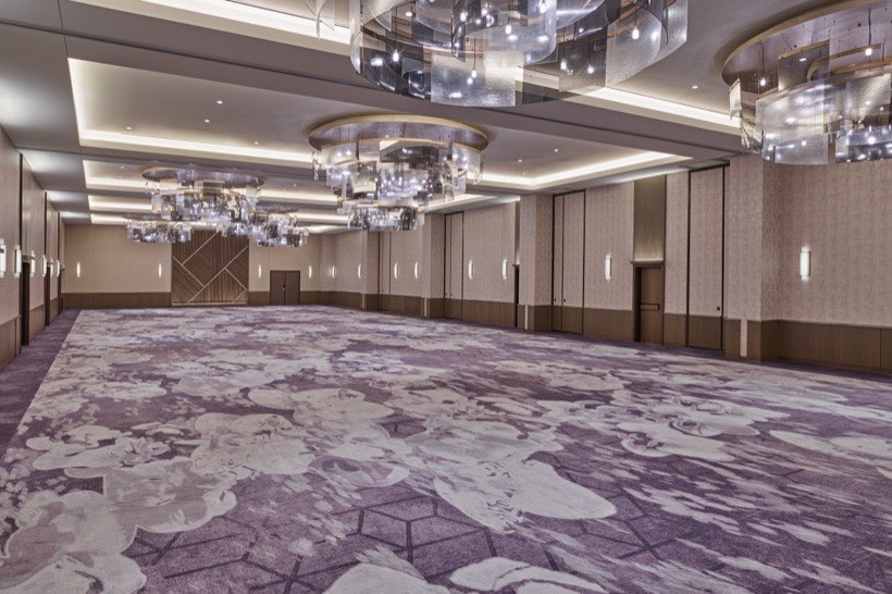 Spacious, empty ballroom with patterned purple carpet, beige walls, modern wall sconces, and large circular glass chandeliers hanging from the ceiling.