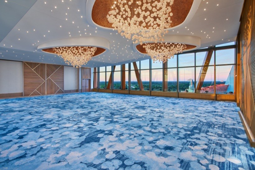 Spacious, modern ballroom with blue patterned carpet, large windows with scenic views, and dramatic chandeliers hanging from the ceiling.