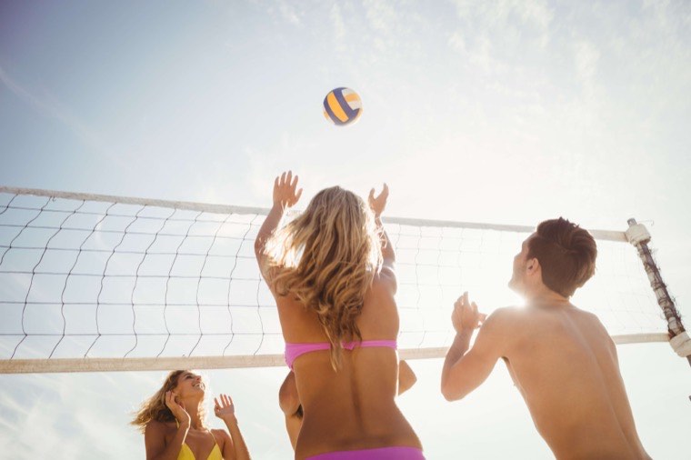 People play beach volleyball under a clear sky. A woman in a pink bikini reaches up toward the ball as others on both sides of the net watch. The sun shines brightly behind them, creating a vibrant, energetic scene.