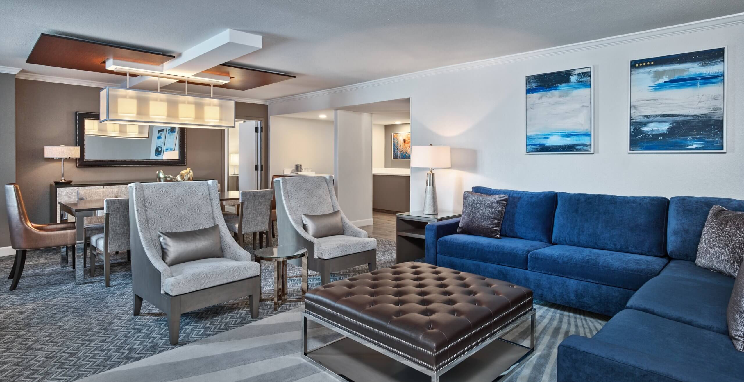 Modern living room with blue sectional sofa, two gray armchairs, tufted leather ottoman, dining table with chairs, and contemporary lighting. Abstract art hangs on the wall above the sofa.