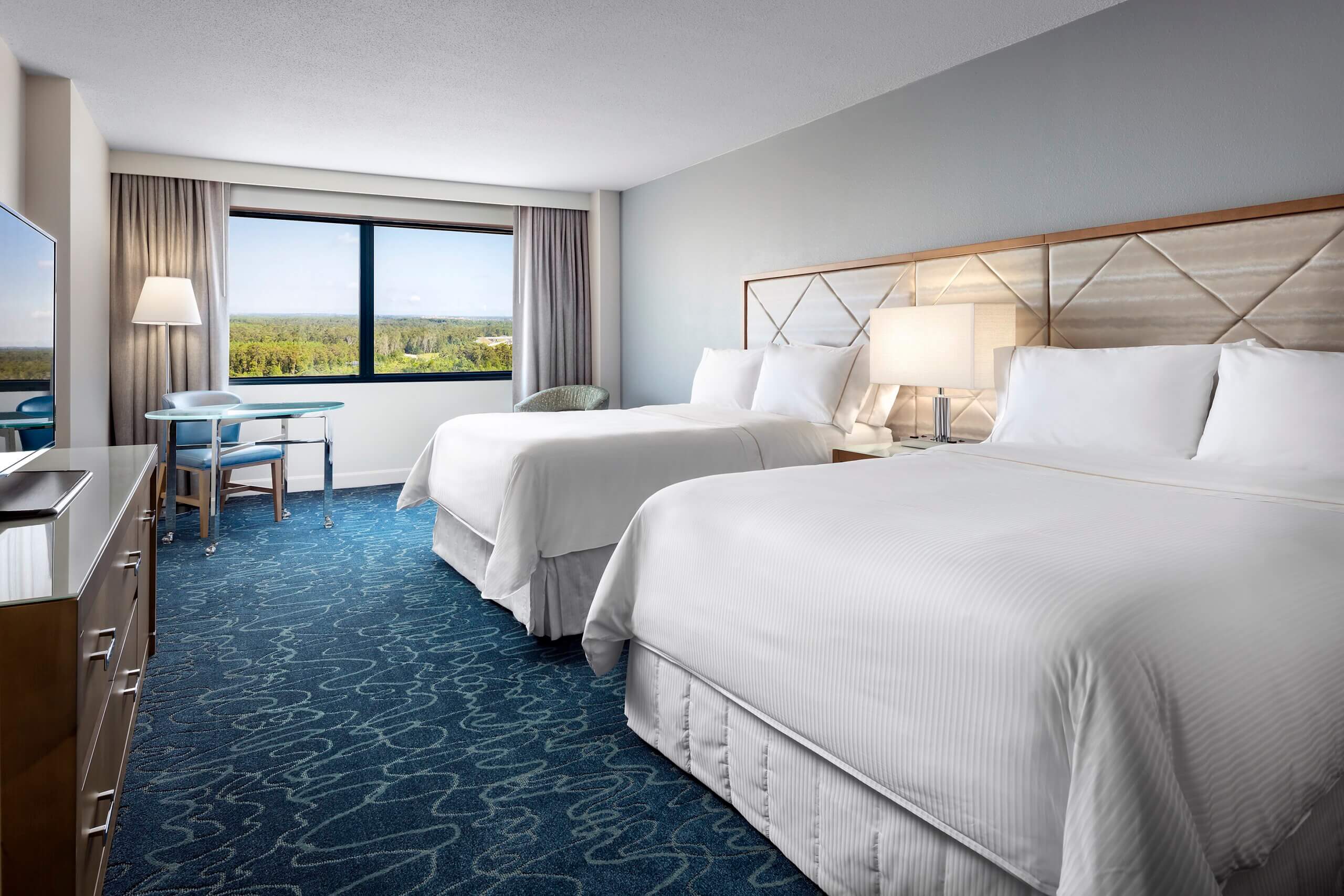 Hotel room with two double beds, white linens, blue carpet, a large window with curtains, a desk, chair, lamp, and a scenic outdoor view. Modern decor with soft lighting and muted color tones.