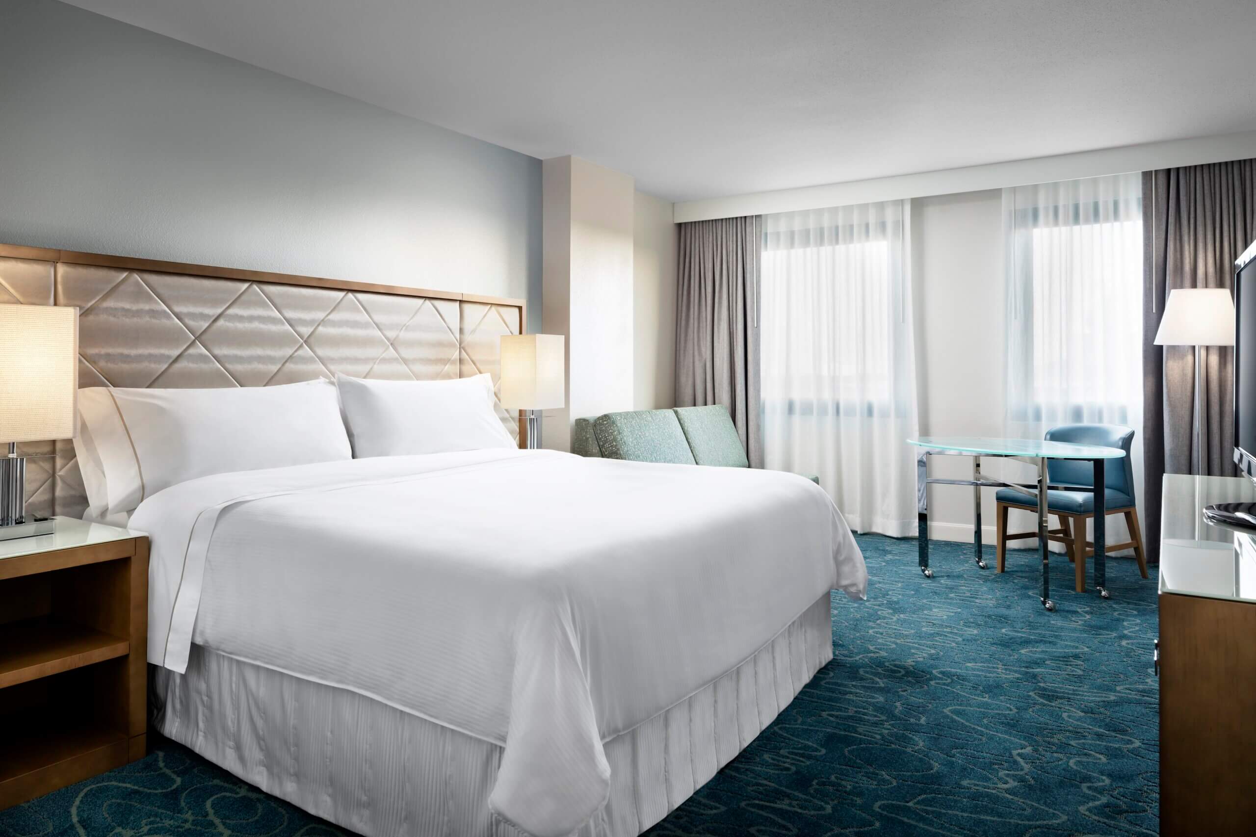 Modern hotel room with a large bed, two bedside lamps, blue carpet, a small couch, a desk with a chair, and two large windows with sheer curtains letting in natural light.