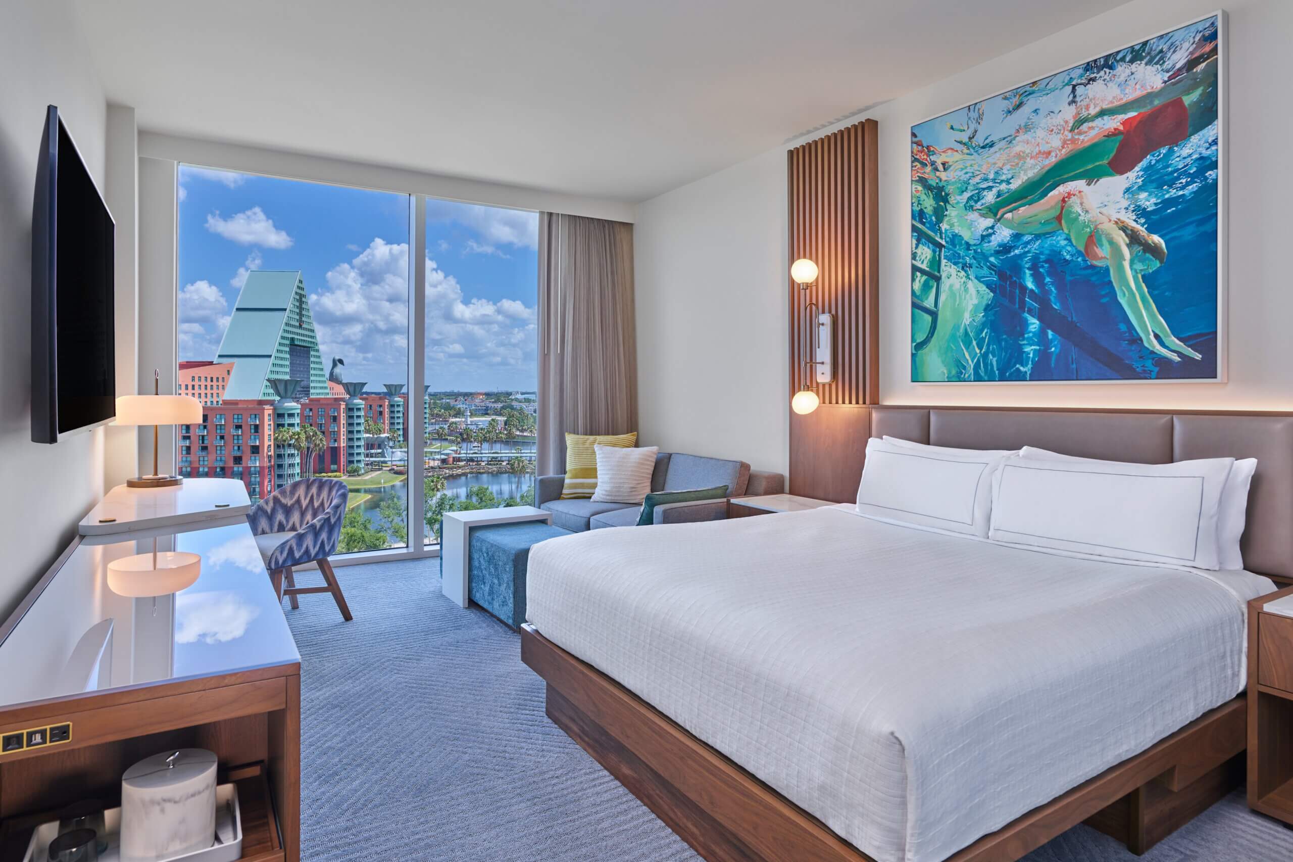 A modern hotel room with a large bed, seating area, desk, and window showing city buildings. Artwork of a swimmer hangs above the bed.