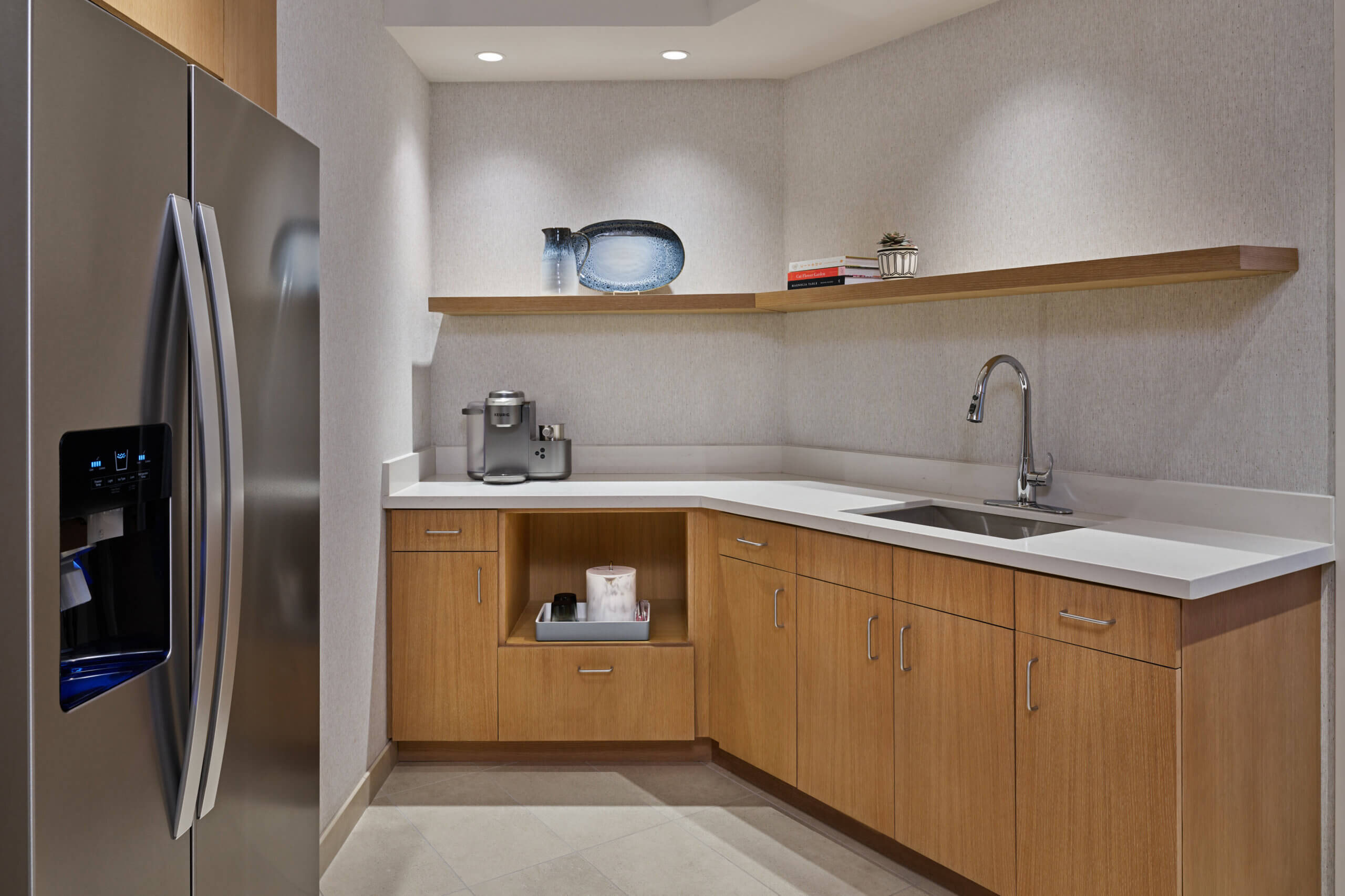 Modern kitchenette with light wood cabinets, stainless steel refrigerator, sink, coffee maker, open shelves with books and decor items, and neutral-colored walls and flooring.