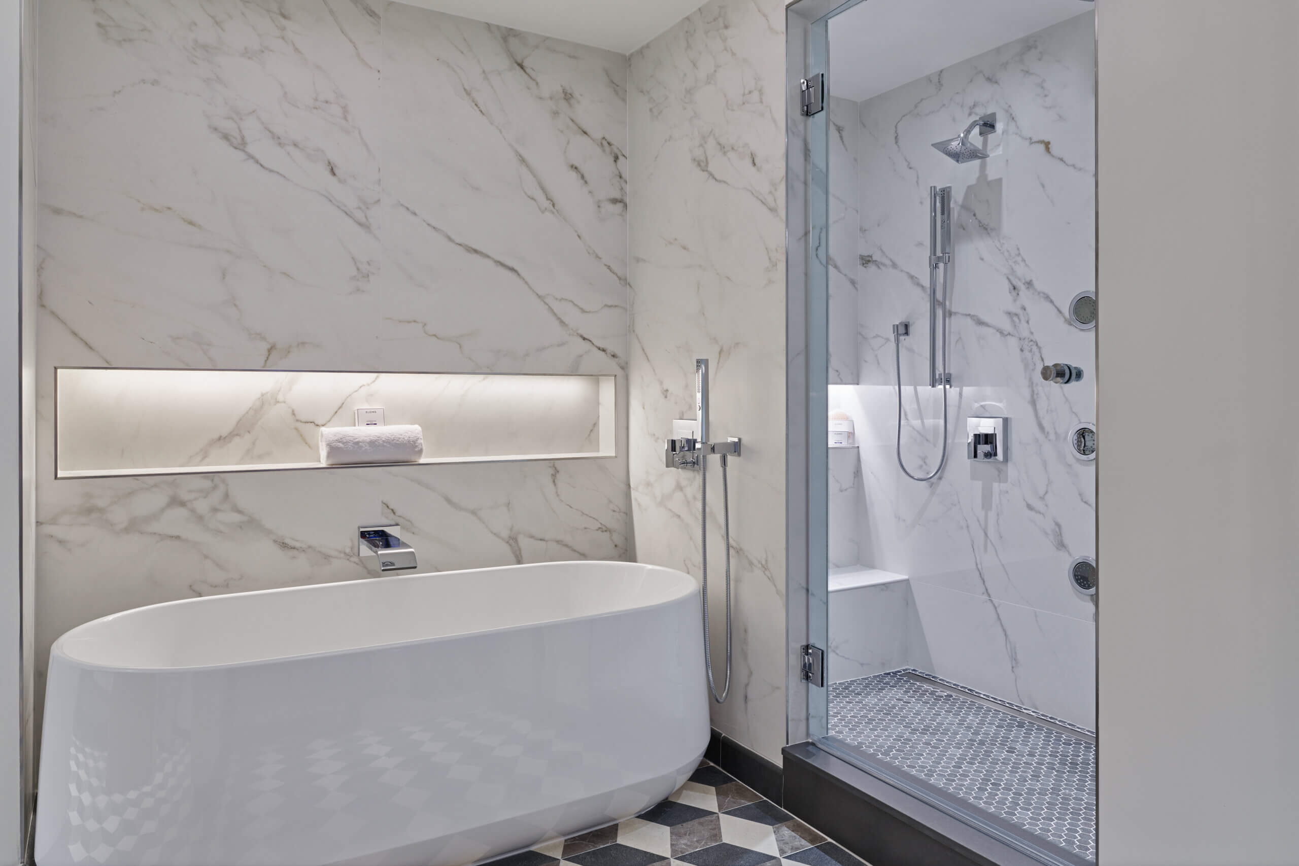 Modern bathroom with a white freestanding tub, marble walls, a built-in shelf with towels, and a glass-enclosed shower featuring a rain showerhead and multiple body jets.
