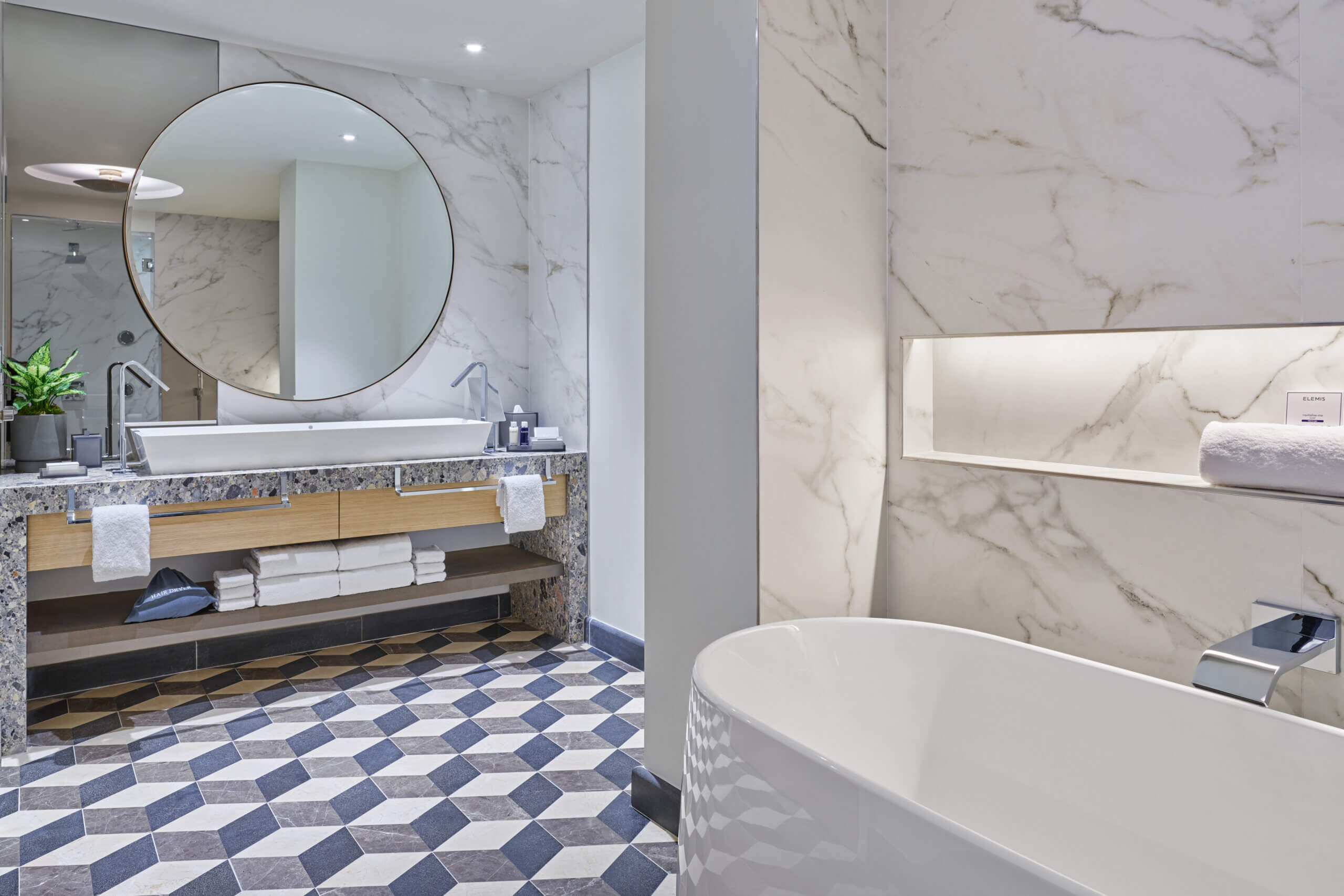 Modern bathroom with marble walls, a freestanding bathtub, geometric tile floor, large round mirror above a sink, open shelving with folded towels, and a small potted plant.