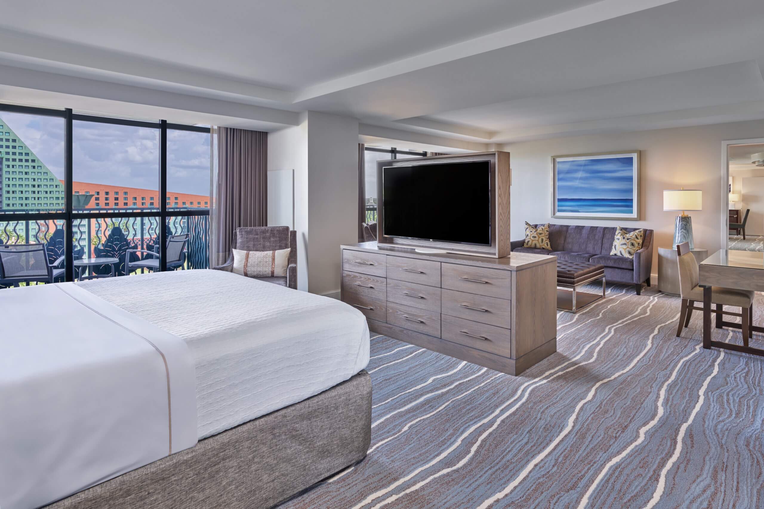 Modern hotel suite with a large bed, flat-screen TV on a dresser, seating area, desk, and balcony with outdoor view. Room has blue and gray tones, patterned carpet, and a framed artwork on the wall.
