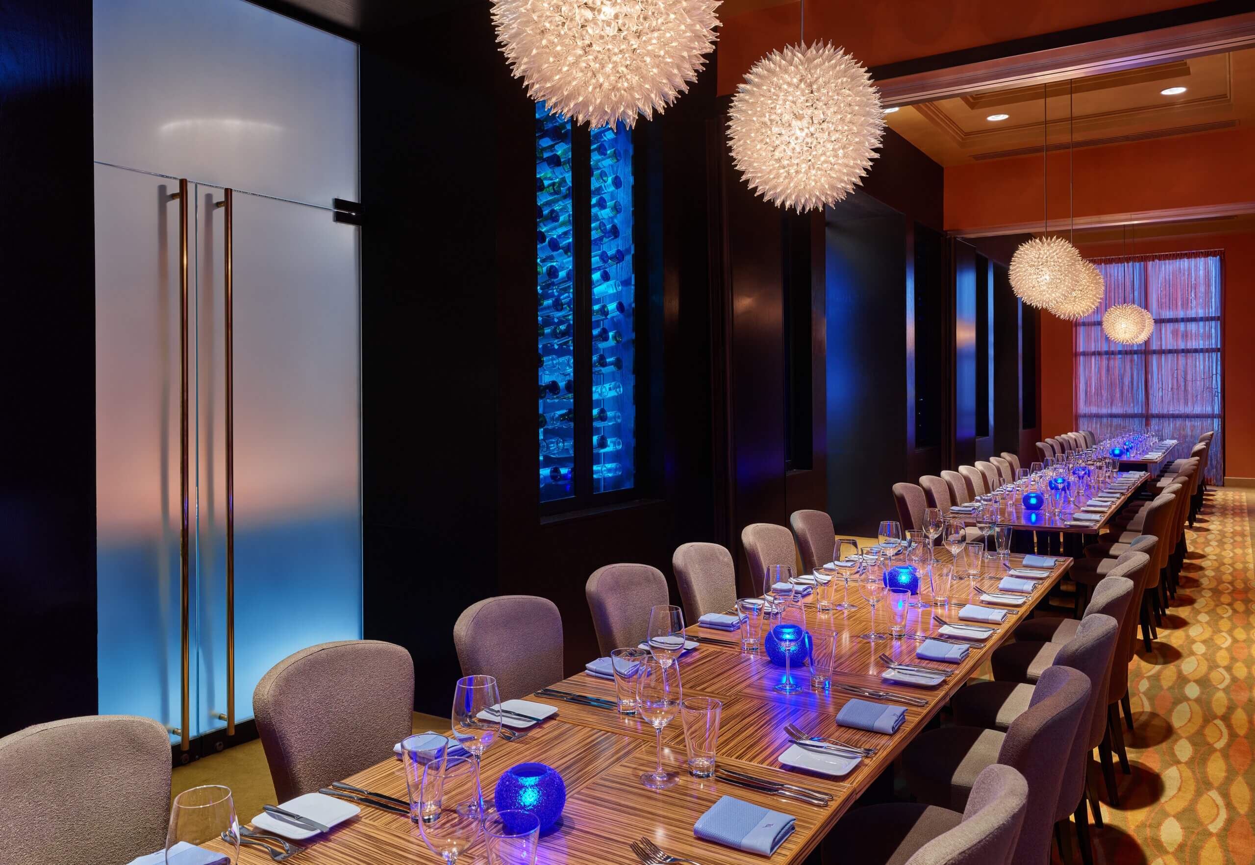 Uma sala de jantar moderna com uma longa mesa de madeira, cadeiras cinzas, castiçais azuis brilhantes, arranjos de mesa elegantes, luminárias pendentes com formato pontiagudo e uma parede de vinhos iluminada em azul ao fundo.