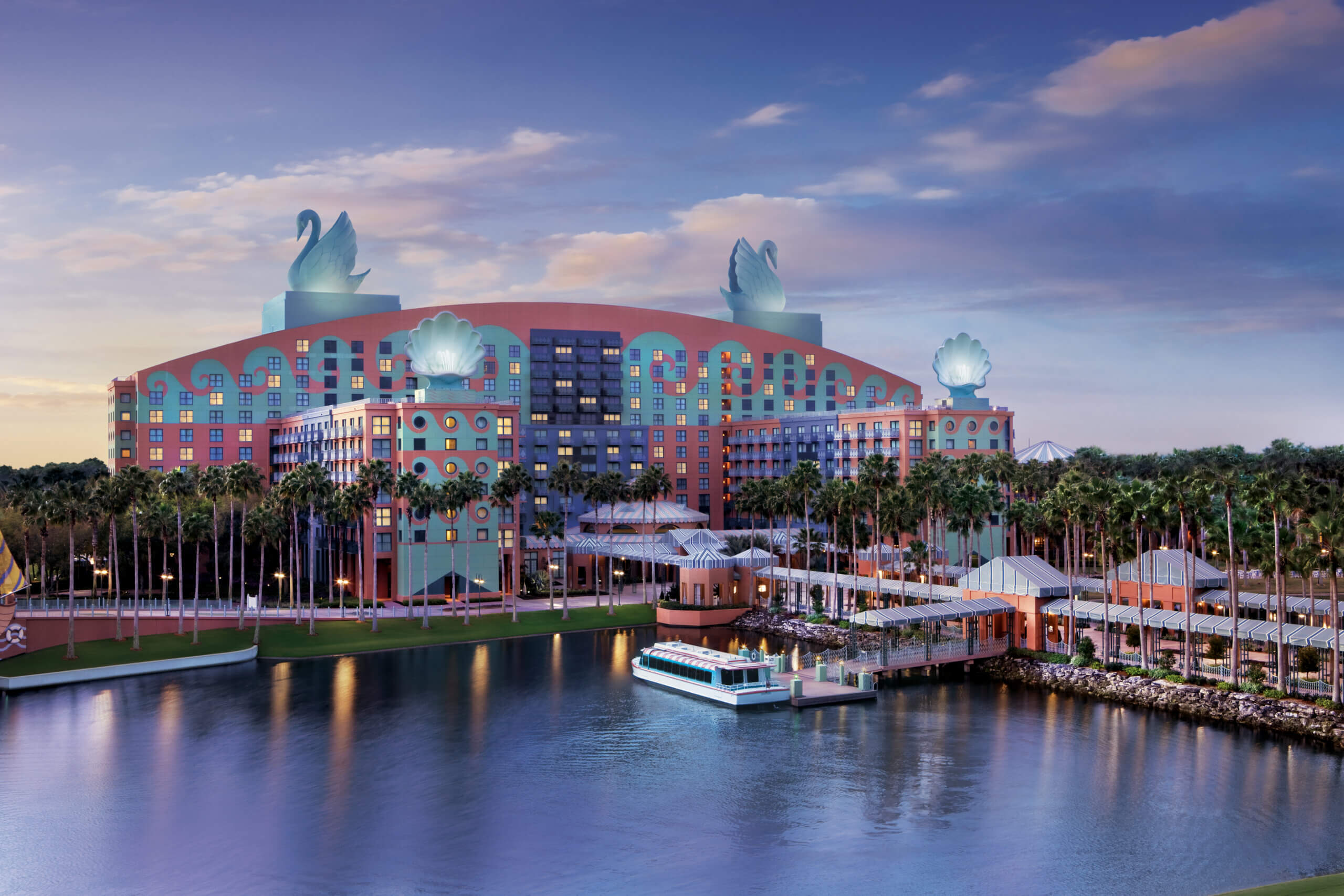 The Disney Swan Hotel at twilight, featuring colorful, post-modern architecture with two large swan statues on the roof, palm trees, a reflecting pool in the foreground, and a small water taxi docked at the pier.