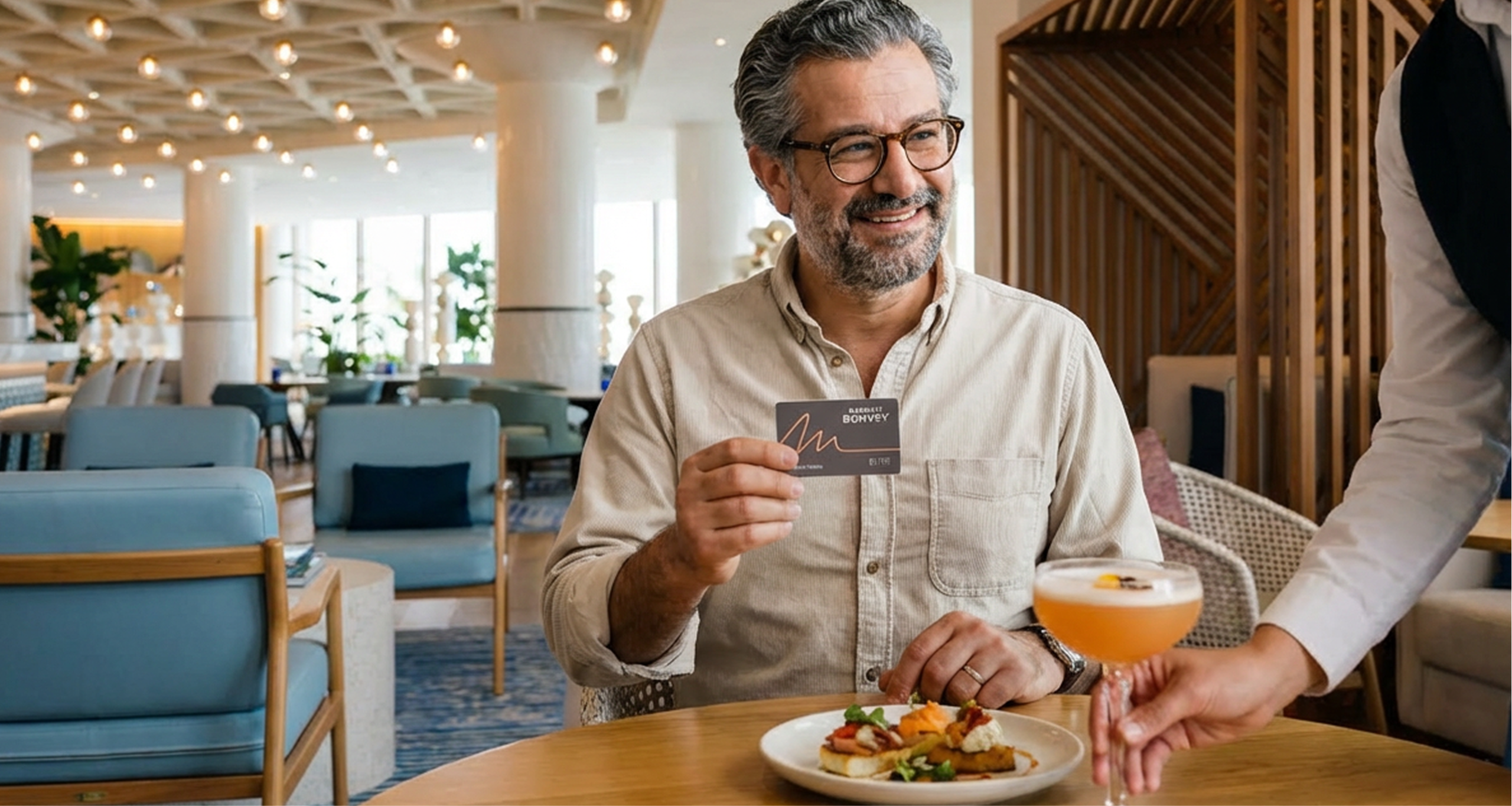 A smiling man sits at a restaurant table, holding up a rewards card. A server places a cocktail on the table next to his plate of food. The setting is bright and modern.