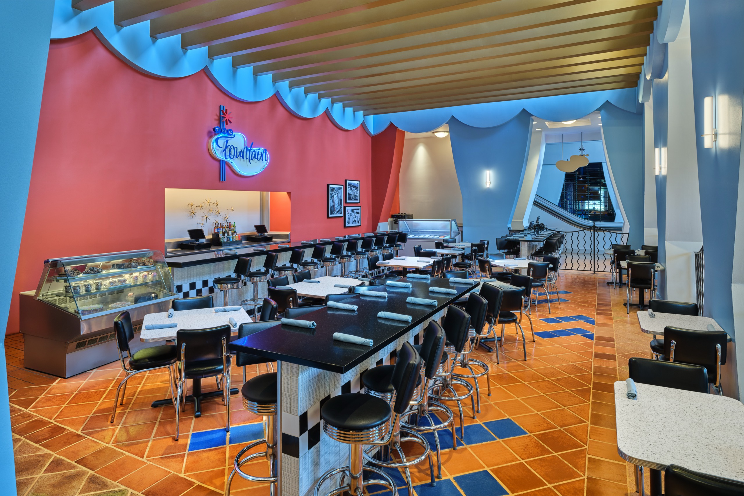A retro-style diner with black stools, checkered tabletops, an orange tile floor, red and blue walls, a counter with a dessert case, and a neon sign reading Florian. Rolled napkins rest on the tables.