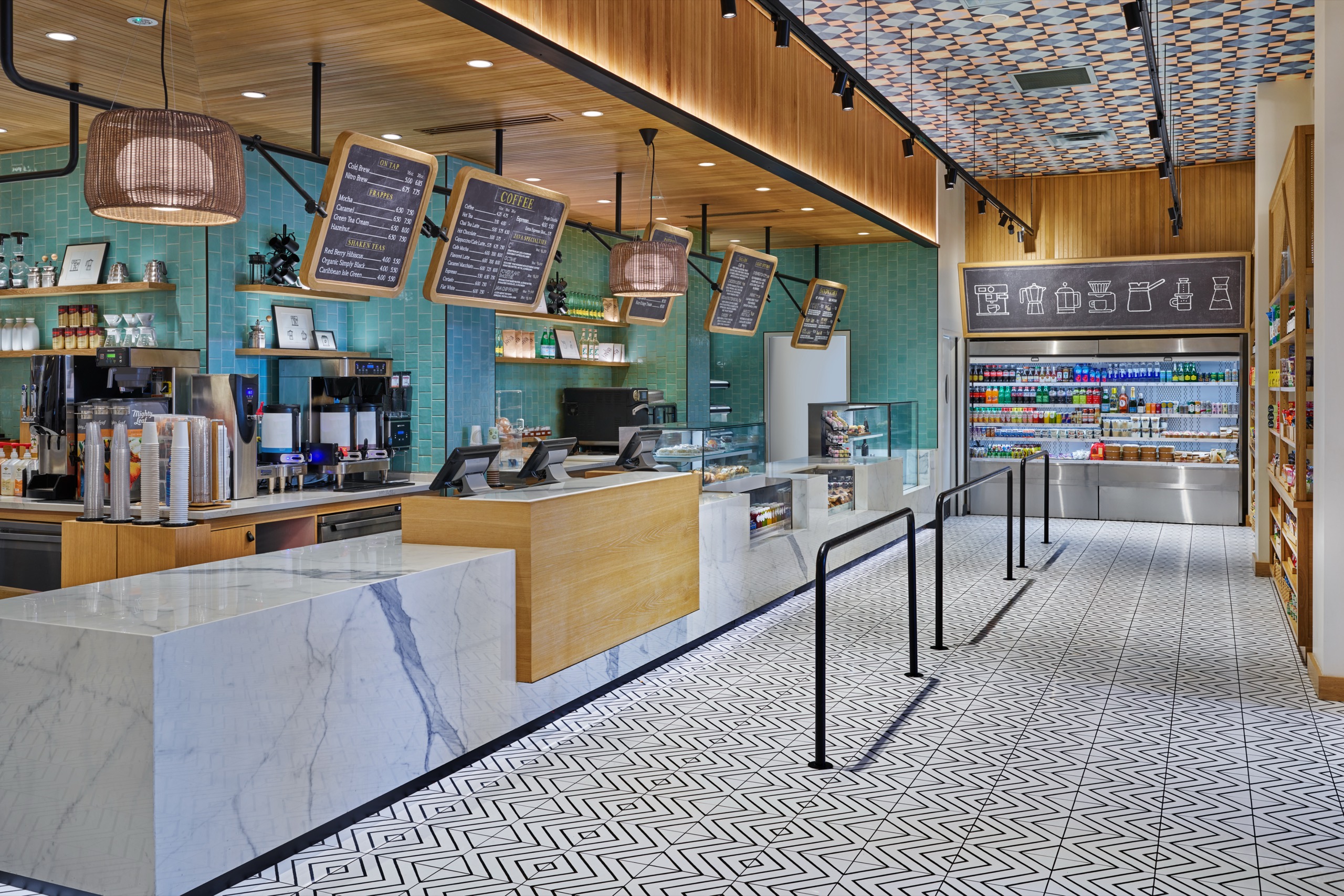 Modern café interior with marble counters, menu boards hanging above, turquoise tiled walls, geometric floor tiles, and a display of drinks in a refrigerated case. Warm lighting and wooden accents create a welcoming atmosphere.