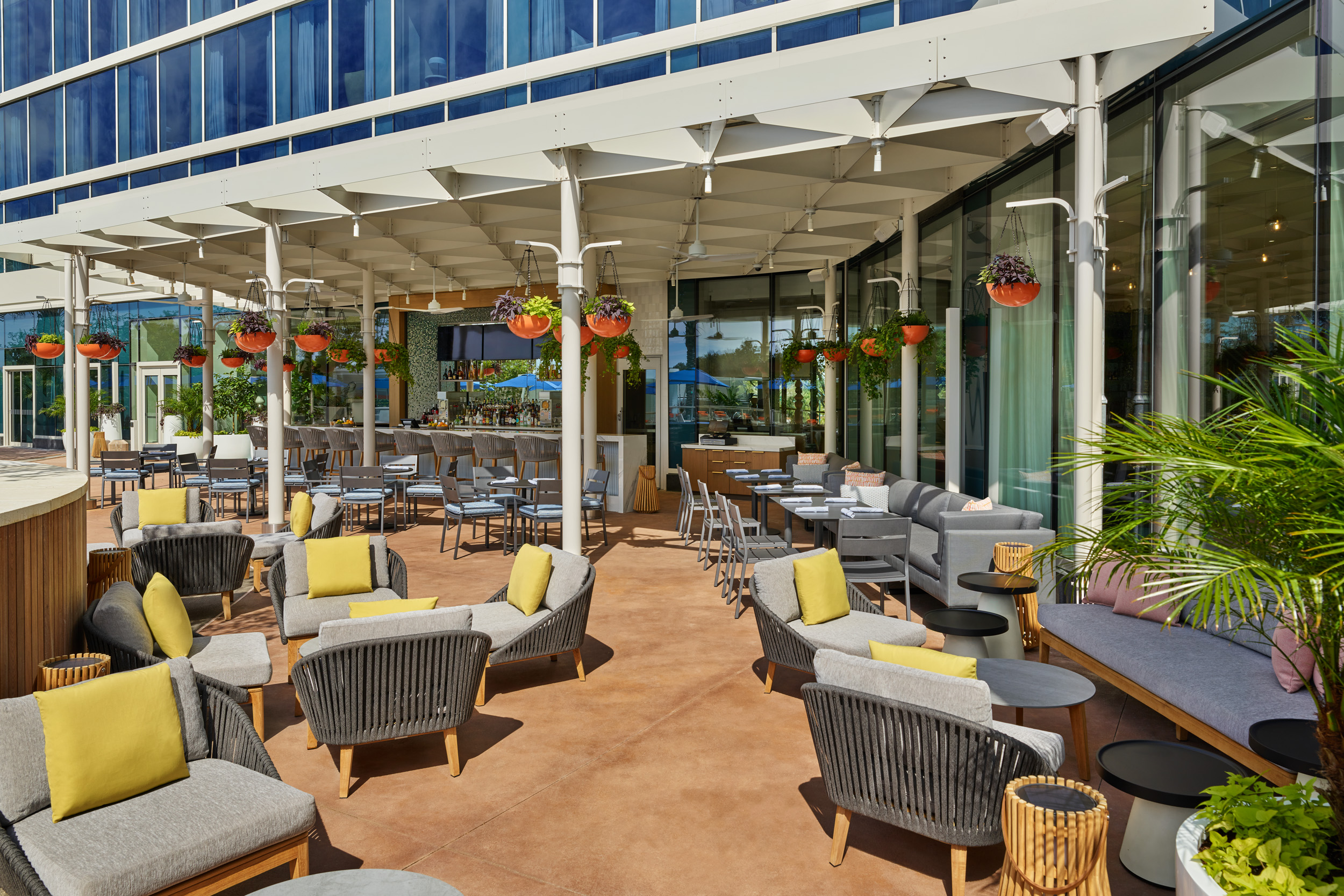 Outdoor patio with modern seating, yellow and gray cushions, round tables, hanging flower pots, and a bar area; glass windows reflect the surroundings. The space is shaded by a white pergola.
