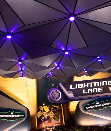 Futuristic ceiling with purple lights above signs for Lightning Lane and Spaceship Earth at an indoor attraction entrance.
