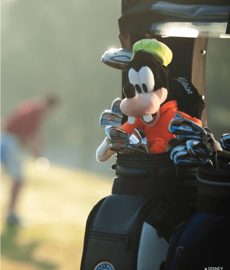 A Goofy plush toy sits in a golf bag filled with clubs, with a golfer blurred in the background on a sunny course.