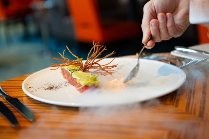 A person uses a spoon to eat an artfully plated gourmet dish on a white plate, with decorative elements and a smoky effect on a wooden table.