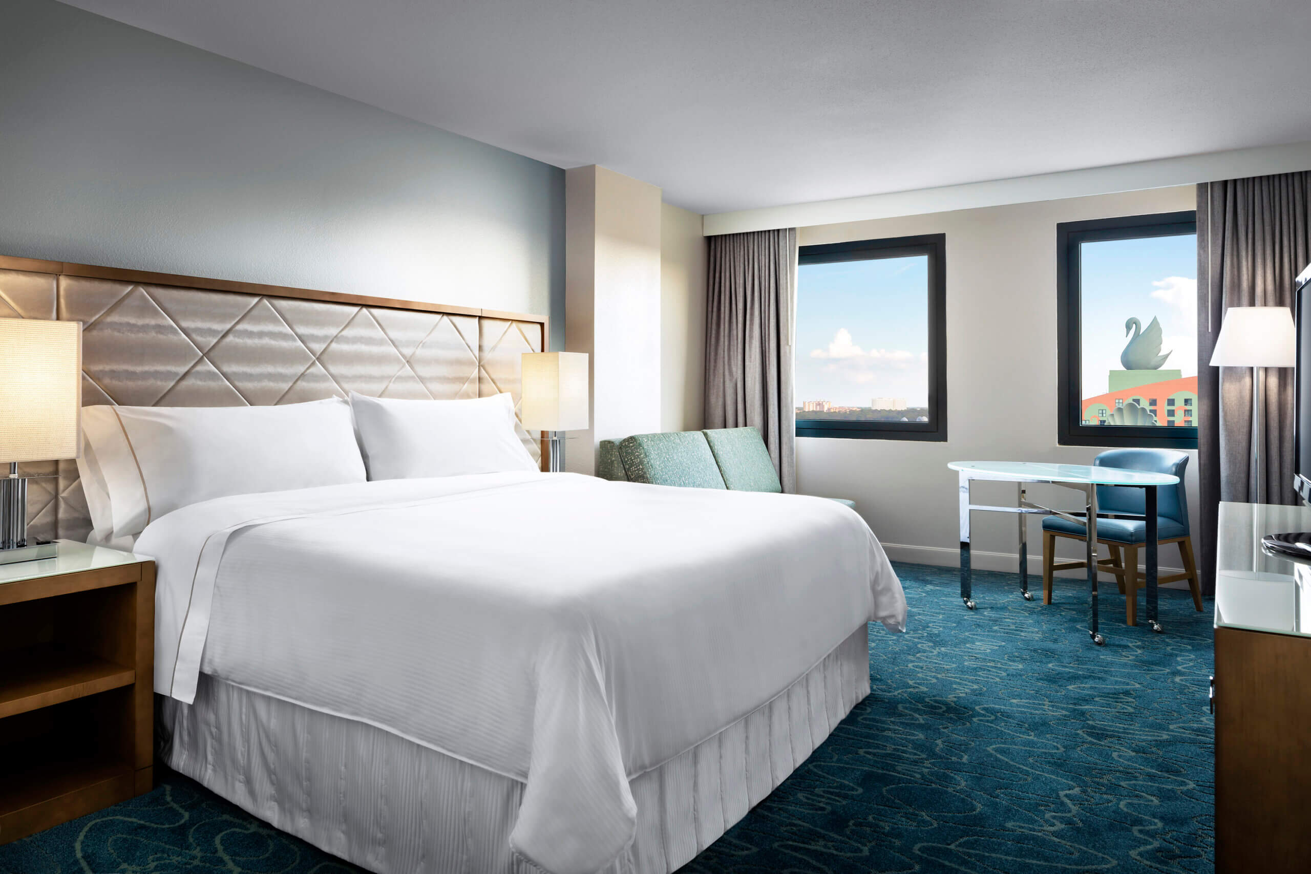 Modern hotel room with a large bed, two bedside lamps, a sofa, a desk, and two windows showing a blue sky and a distant swan statue. The room features blue carpet and neutral walls.