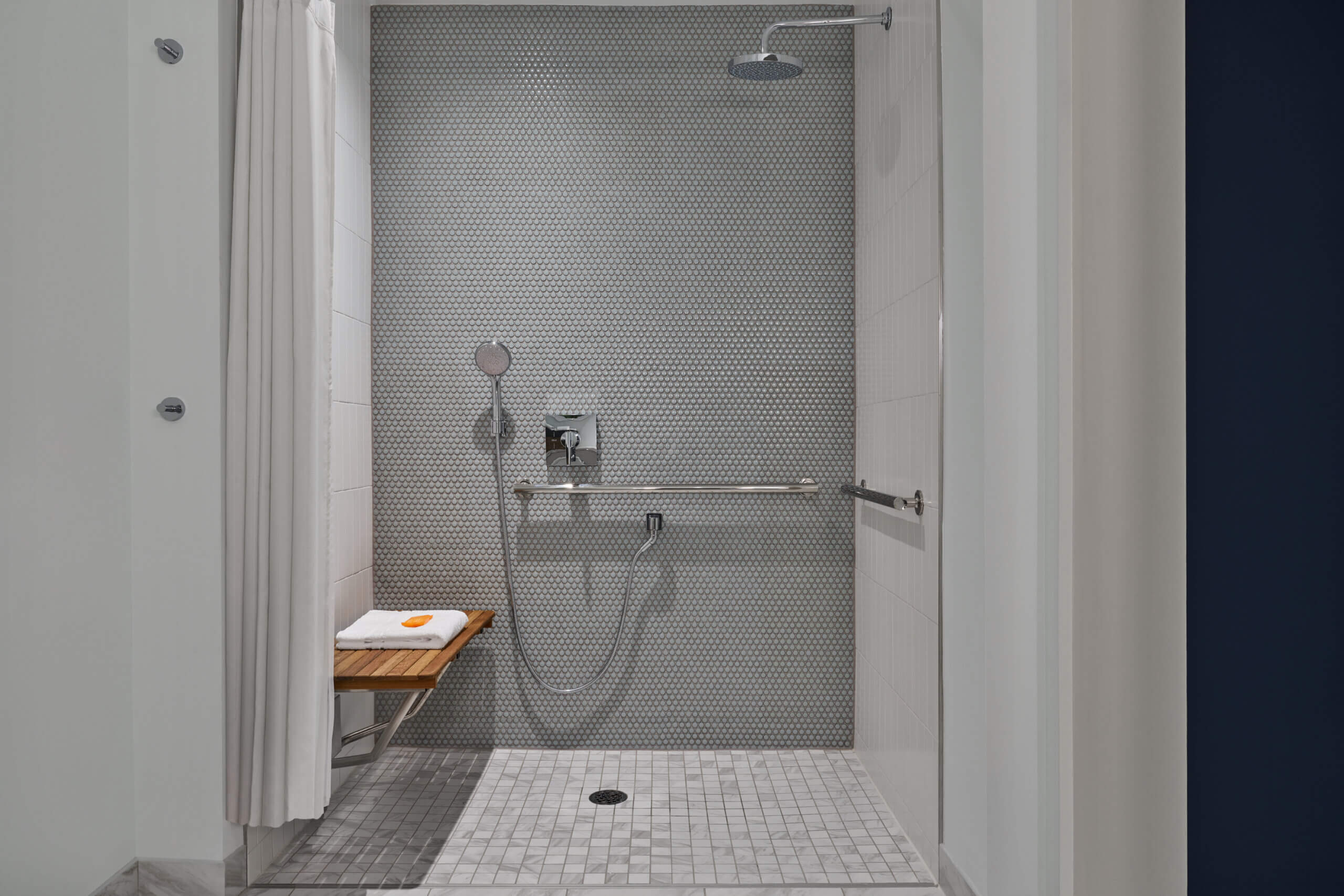 Accessible walk-in shower with a mounted showerhead, handheld shower, grab bar, and a fold-down wooden bench with a towel on it. Light gray tile lines the floor and wall. A white shower curtain is partially open.