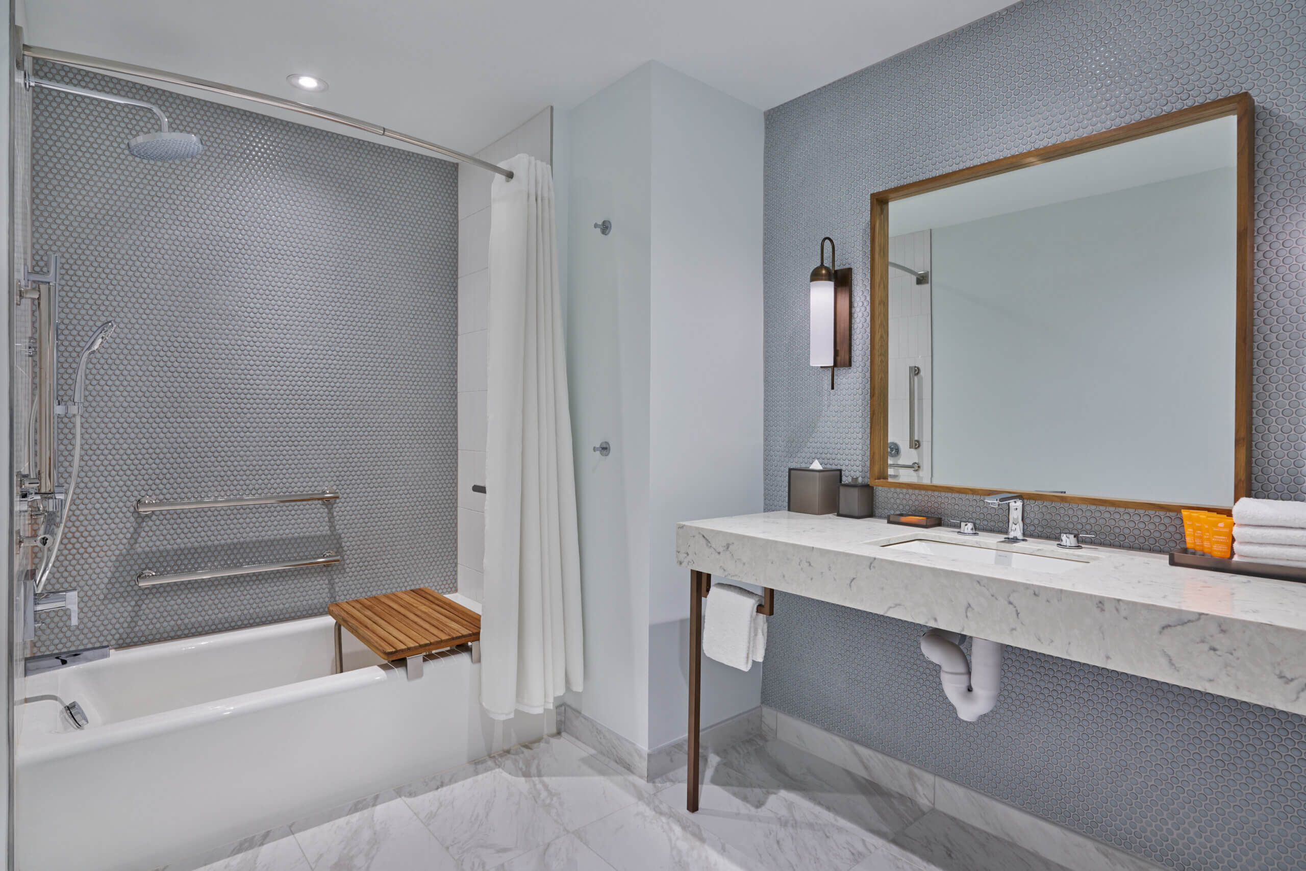 Modern bathroom with a bathtub featuring a wooden seat, grab bars, a showerhead, and a white curtain. A marble countertop holds towels and toiletries, with a mirror and wall sconce above.