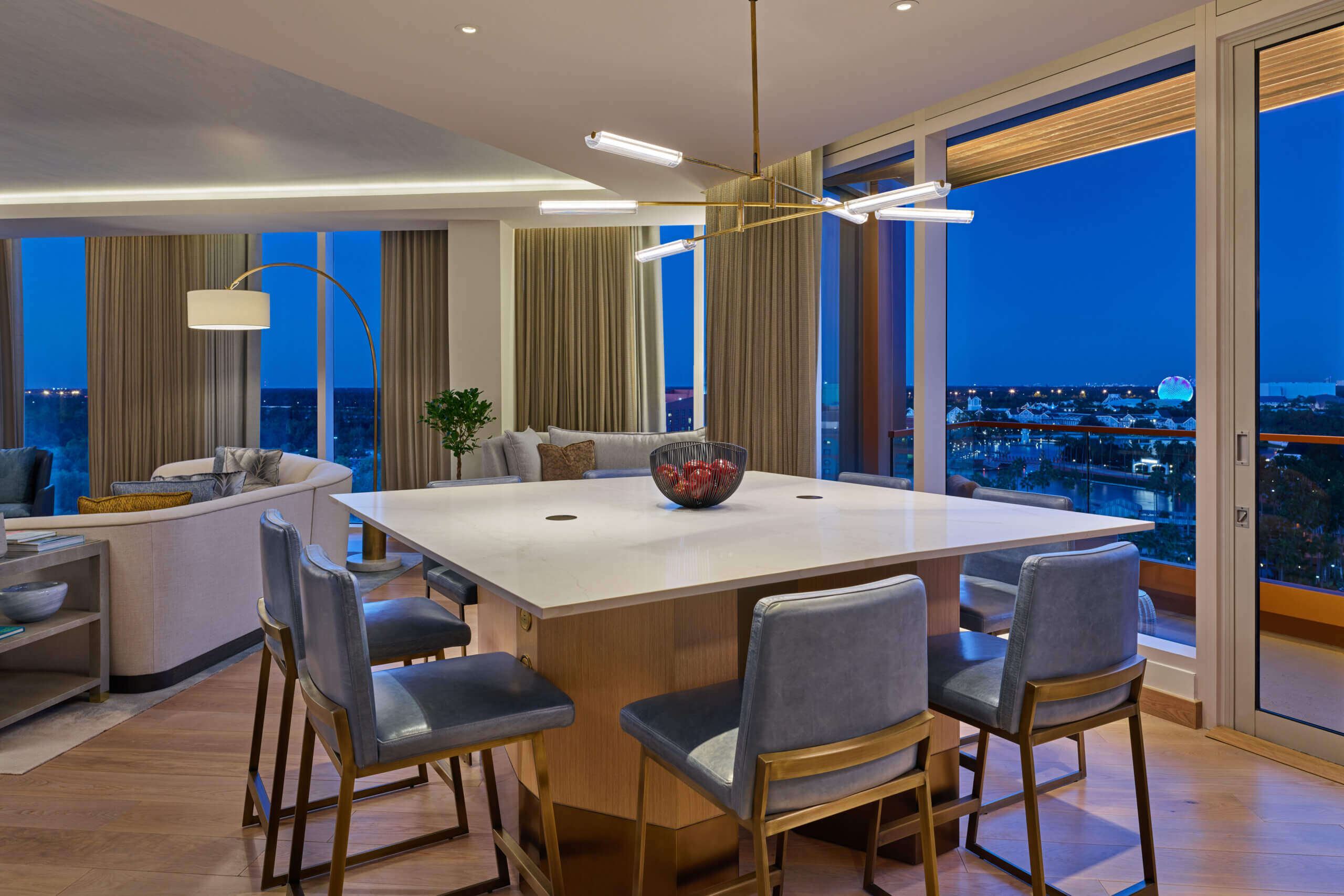 Modern dining area with a large white table, four gray chairs, and a bowl of red apples. Floor-to-ceiling windows reveal a cityscape at dusk. A stylish chandelier and cozy living area are visible in the background.